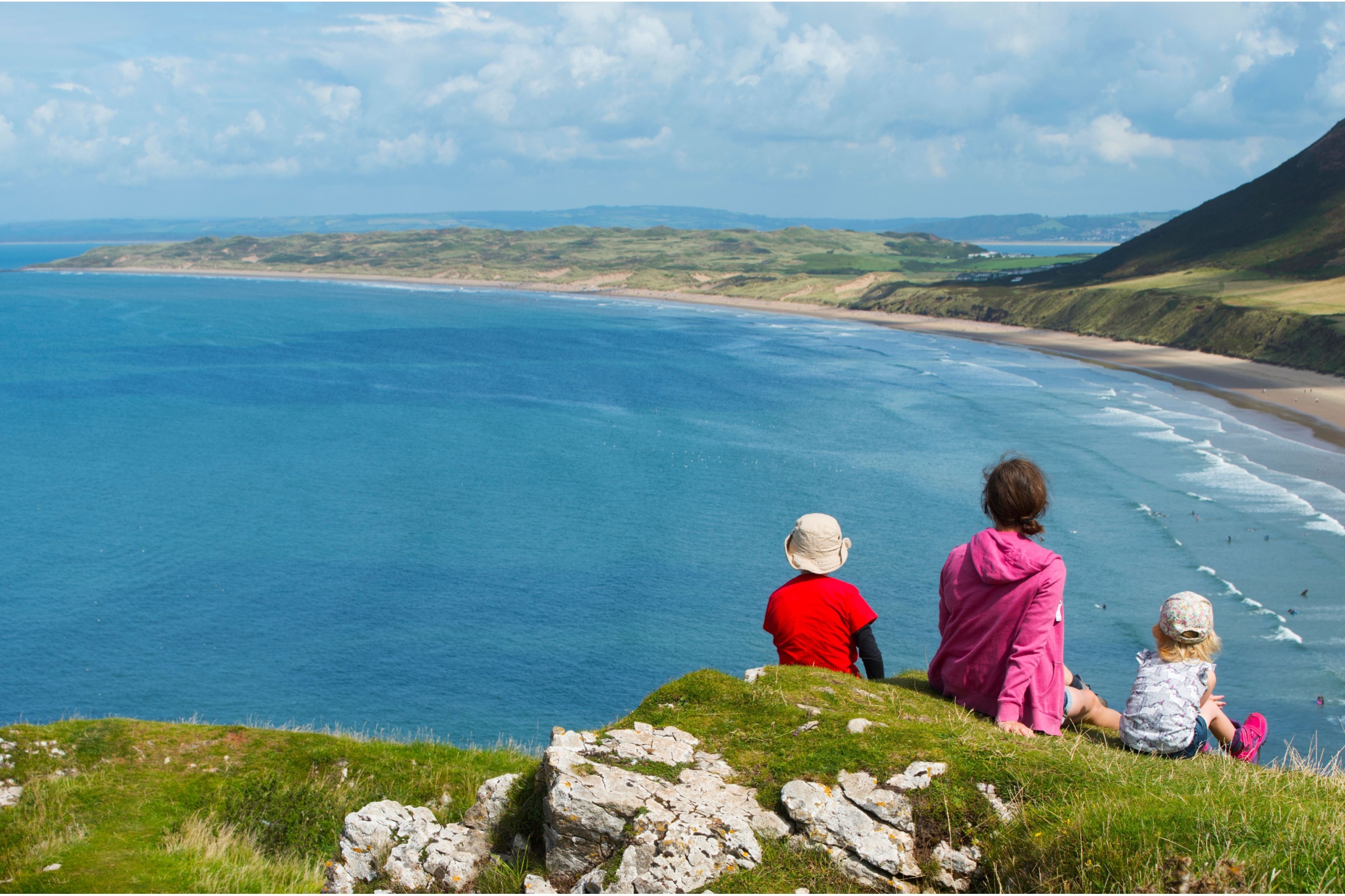 A family sitting on a clifftop overlooking a beach.